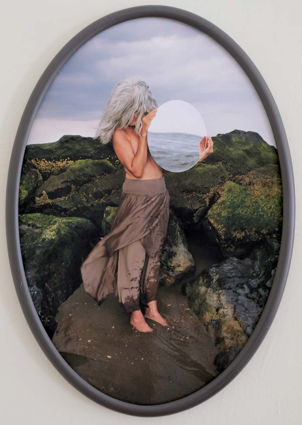 Long grey-haired woman standing in mud on rocky shore holding a circular mirror reflecting the ocean